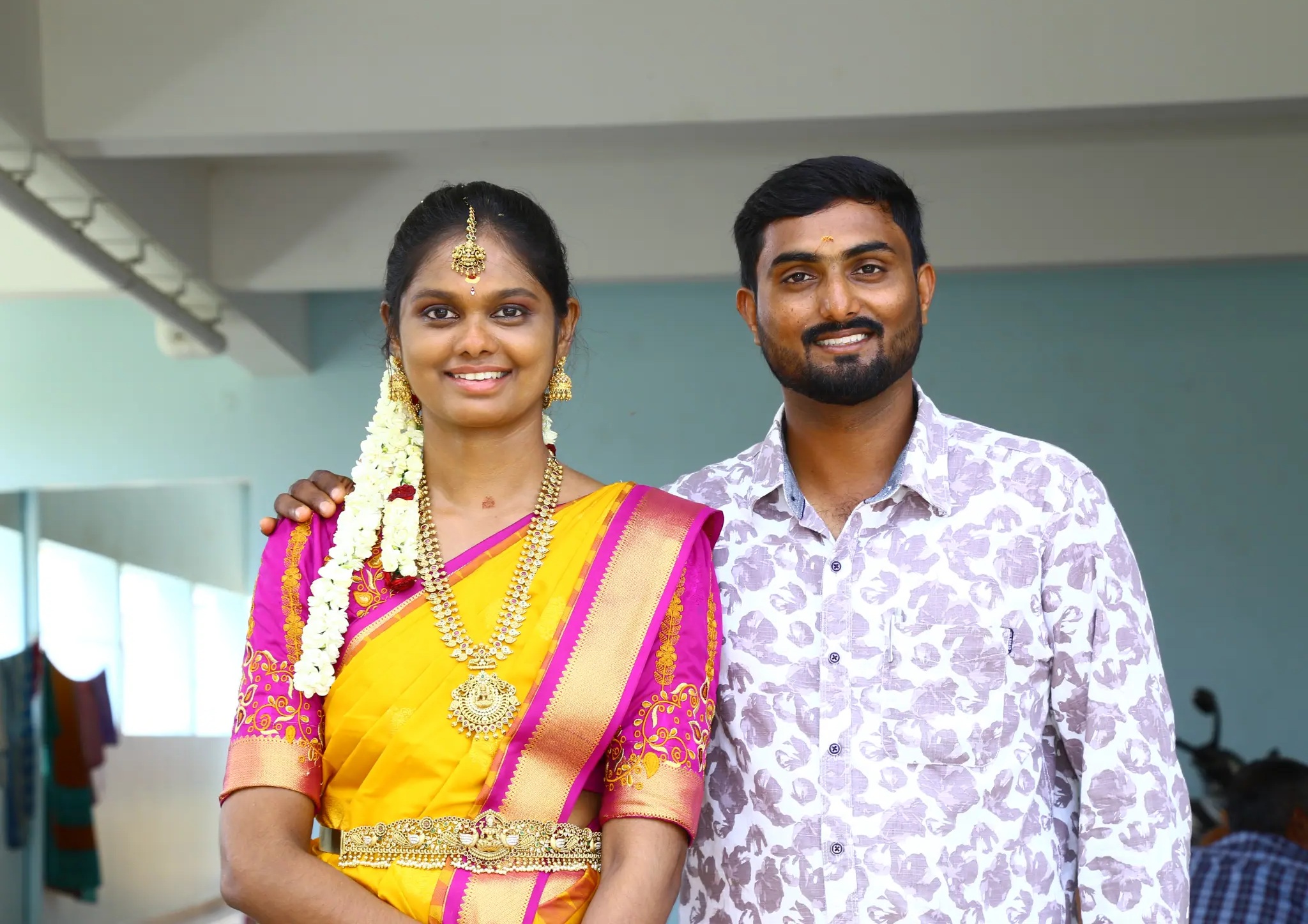 Naveen and Ramya together, Ramya wearing a yellow and magenta sari and Naveen wearing a light patterned shirt